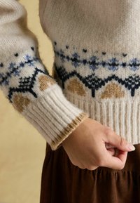 Strickpullover mit cremefarbenem Grund, mehrfarbigem geometrischem Muster in Blau und Braun, gerippten Bündchen und einer weichen, strukturierten Oberfläche.