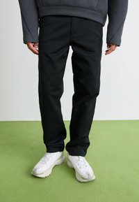 Persoon in zwarte broek, witte sneakers en een donkergrijze lange mouwtop die op een lichtgroene vloer staat tegen een witte muur.
