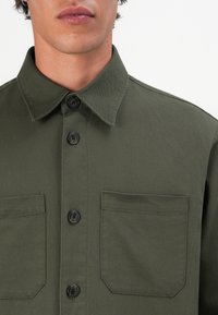 Hombre vestido con una camisa de botones color verde oliva, con dos bolsillos en el pecho, cuello y botones oscuros, contra un fondo blanco.