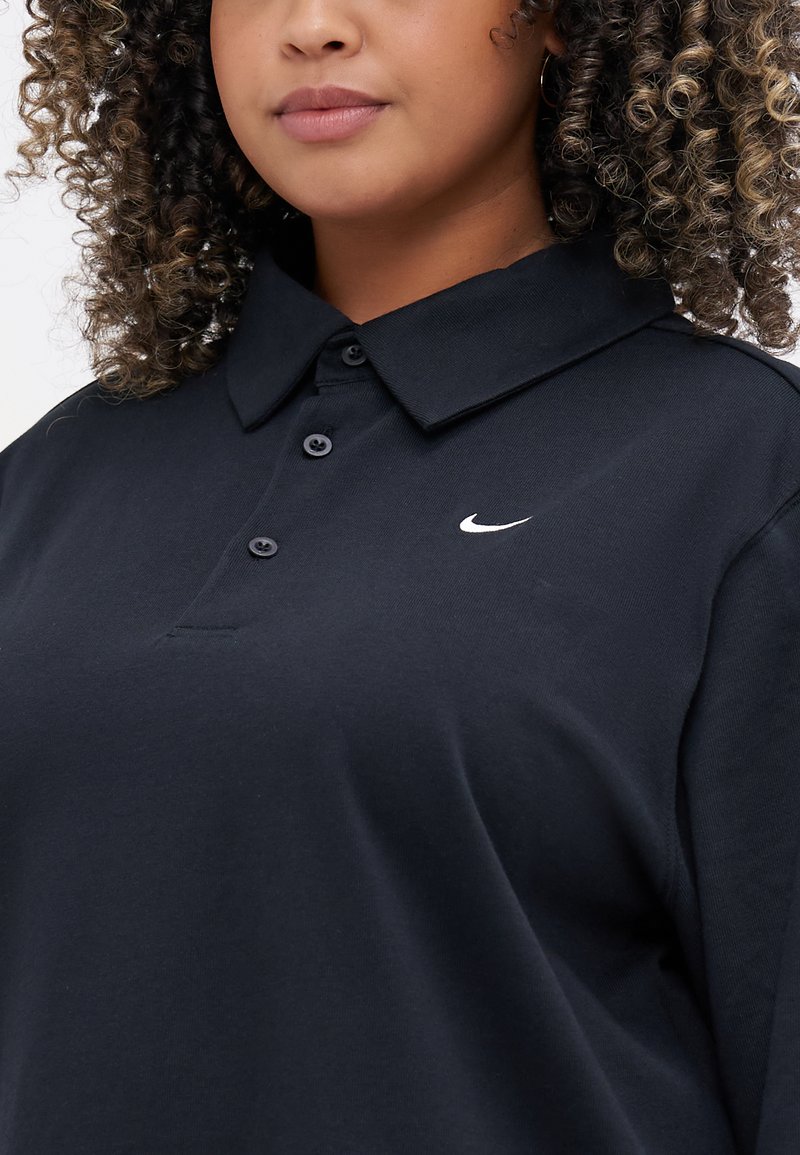 Personne aux cheveux bouclés portant un polo noir Nike à trois boutons avec un logo swoosh blanc sur la poitrine.