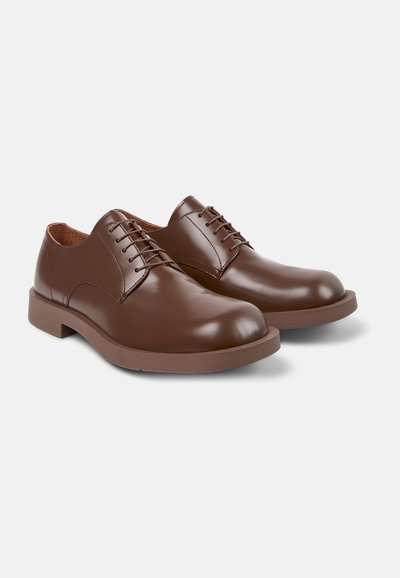 Scarpe in pelle marrone con una texture liscia, punta arrotondata e design con lacci. Presentano una suola in gomma marrone solida e minimalista.