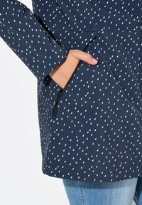 Giacca impermeabile navale con motivo a goccia bianca, dotata di tasca con zip e design leggermente aderente. Tessuto morbido e liscio.