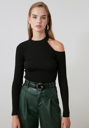 Jeune femme portant un haut côtelé noir avec une découpe à une épaule et un pantalon en cuir vert foncé taille haute avec ceinture noire.