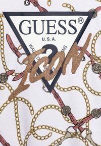 T-shirt bianco in tessuto con stampa di catena dorata e fascia rosa. Presenta un'etichetta triangolare nera con la scritta "GUESS" e il testo "ICON" glitterato in bronzo.