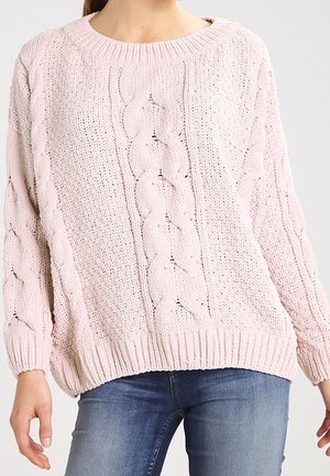 Femme portant un pull en maille torsadée rose clair et un jean bleu, debout devant un fond clair uni.