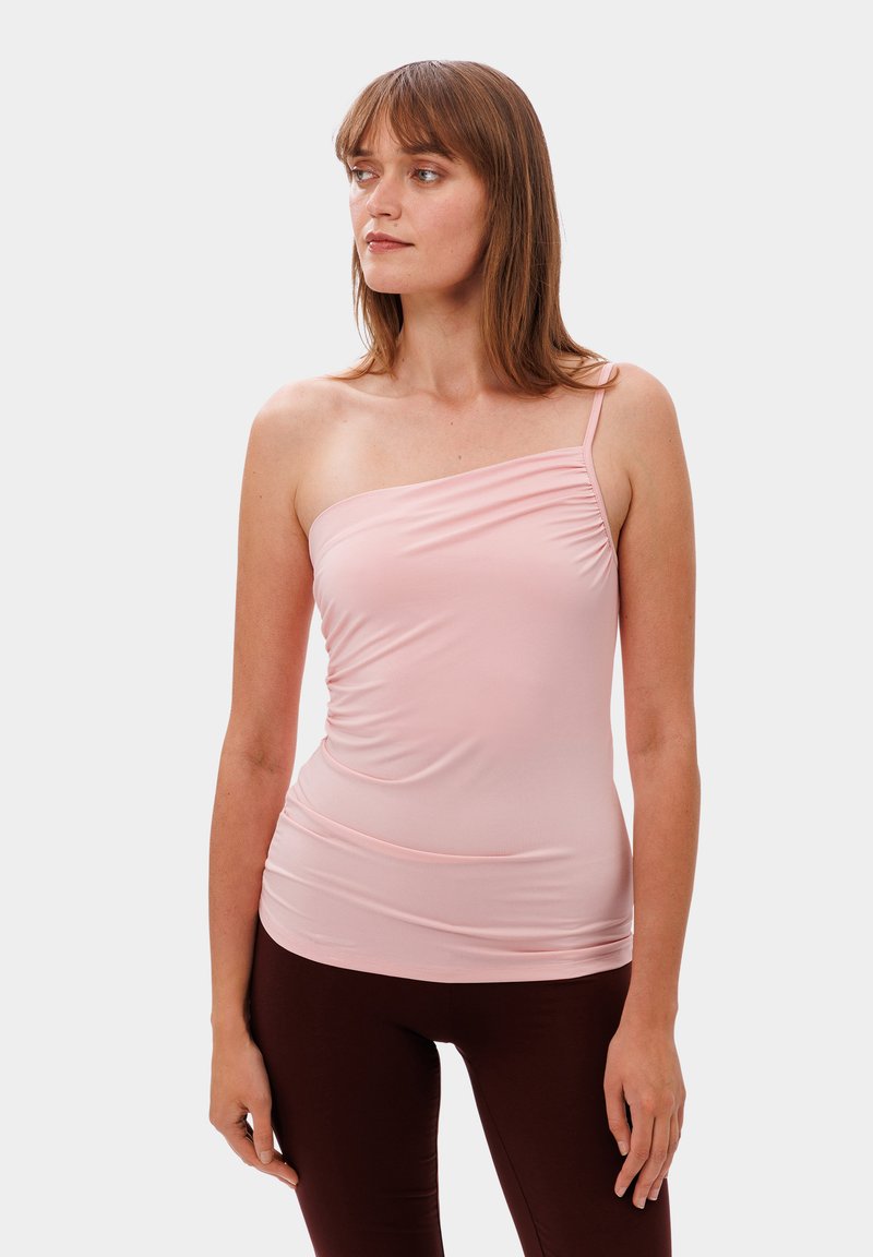 Světle růžový top na jedno rameno z elastické tkaniny, který má na přední straně řasený design a hladkou strukturu, kombinovaný s hnědými legínami.