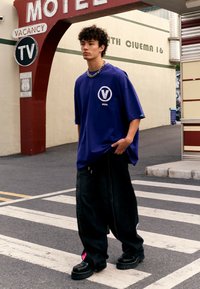 Una camiseta morada de gran tamaño presenta un logotipo circular en blanco. Se combina con unos jeans negros anchos y zapatos negros voluminosos con acentos rosas.