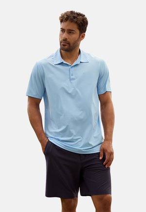 Hombre con camisa polo azul claro y pantalones cortos azul marino oscuro, de pie con las manos en los bolsillos, mirando ligeramente hacia un lado contra un fondo liso.