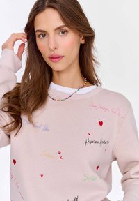 Světle růžová mikina s kontrastním bílým výstřihem; obsahuje barevný ručně psaný text a malé grafiky srdíček po celém povrchu.