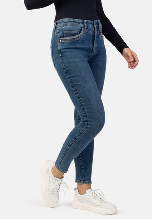 Dunkle Jeans im Skinny-Fit, hoch tailliert mit fünf Taschen, Metallakzenten und glatter Textur; kombiniert mit weißen Sneakern.