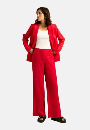 Red blazer and matching wide-leg pants worn over a white tank top. The outfit features a smooth texture and minimalistic design. Black sandals complete the look.