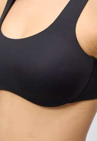 Artimo asmuo dėvi juodą, be siūlių, bevielę liemenėlę su plačiomis petnešėlėmis ir lygiu audiniu.