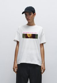 Bílá bavlněná tričko s grafickým potiskem ženské tváře a textem v růžové barvě, kombinované s tmavými kalhotami a černou kšiltovkou.