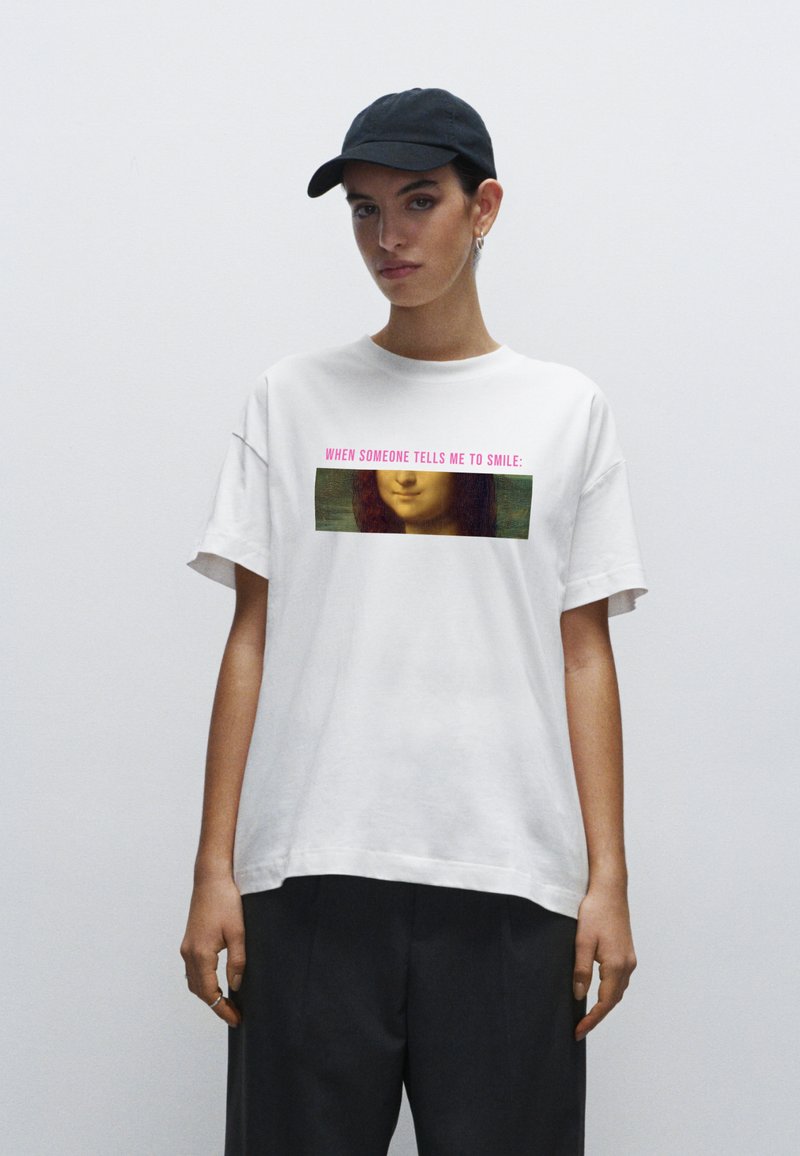 Bílá bavlněná tričko s grafickým potiskem ženské tváře a textem v růžové barvě, kombinované s tmavými kalhotami a černou kšiltovkou.