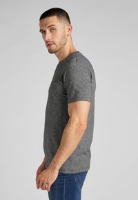 Grå crew-neck t-shirt lavet af struktureret stof, med kortærmer og en brystlomme, parret med blå jeans. Sideprofil udsigt.
