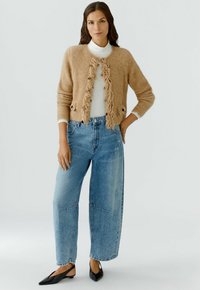 Beige gebreide cardigan met franjes, over een witte coltrui, gecombineerd met lichtblauwe hoge taille wijde jeans en zwarte puntige flats.