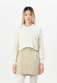 Fiatal nő egyenes hajjal, krém színű, fonott mintájú, rövid pulóverben, bézs szoknyával és fehér leggings-szel, egy sima háttér előtt.