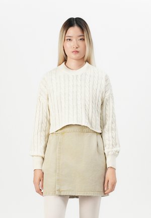 Young woman with straight hair wearing a cream cable-knit cropped sweater over a beige skirt and white leggings against a plain background.