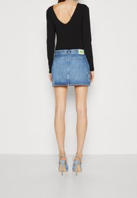 Svart långärmad bodysuit med djup V-rygg, i kombination med en blå denimminikjol med fickor och en gul tagg vid midjan.