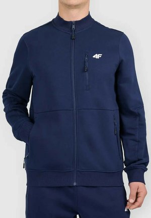 Man draagt een marineblauwe jas met ritssluiting, drie zakken met rits en een wit 4F-logo op de borst.