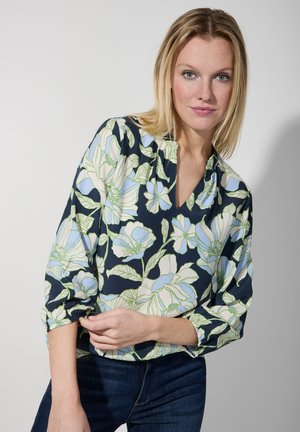 Blouse à motifs floraux sur fond marine, ornée de grandes fleurs blanches et bleues claires ainsi que de feuilles vertes. Décolleté en V et manches froncées.