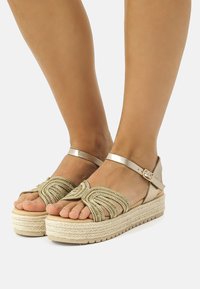 Kultaisten espadrille-sandaalien yläosa on punottu, ja niissä on nilkkaremmitys sekä kudottu juuttipohja. Littee korko, avokärkinen tyyli.