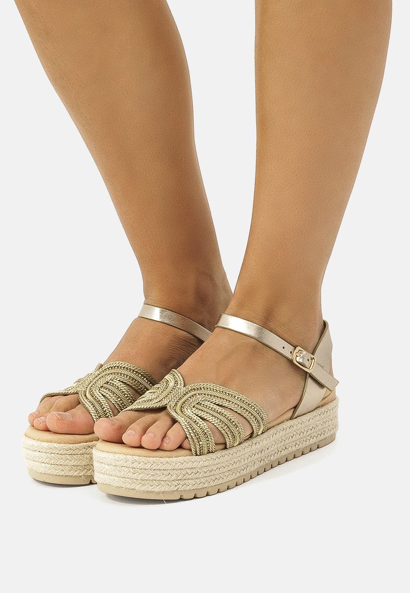 Kultaisten espadrille-sandaalien yläosa on punottu, ja niissä on nilkkaremmitys sekä kudottu juuttipohja. Littee korko, avokärkinen tyyli.