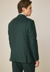 Sacou din lână de un verde închis, cu umeri structurați, rever în formă de săgeată și un singur slit. Prezintă manșete cu buton și o textură netedă.