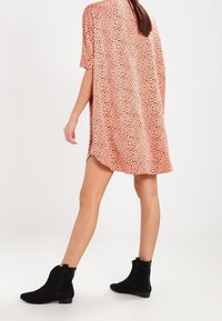 Femme portant une robe rose ample avec des pois noirs et des bottines noires, se tenant avec une jambe légèrement en avant sur un fond blanc.