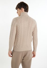 Beige, kabelhálós pulóver magas gallérral, textúrált mintákkal és bordázott aljjal. Világos színű nadrággal viselve.