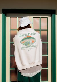 Persona che indossa un cappello a falda bianco e una felpa Reebok bianca con grafica di un campo da tennis, in piedi davanti a una porta a vetri con modanature verdi e pannelli multipli.