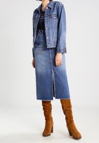 Denim jacket with a fitted design, paired with a matching denim skirt featuring a front slit. Complemented by knee-high tan suede boots.