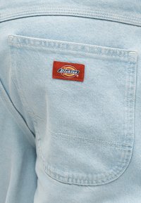 Πίσω τσέπη από ανοιχτό μπλε τζιν με ραμμένη κοντά στο επάνω κέντρο της τσέπης ετικέτα της μάρκας Dickies σε κόκκινο και κίτρινο χρώμα.