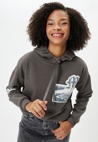 Grijze hoodie met een voorzak, trekkoord bij de hals en een grafische print. Model draagt het met donkere jeans.