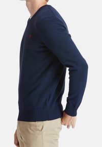 Tmavě modrý pletený svetr s V-výstřihem, žebrovanými manžetami a lemem. Malý červený detail loga na hrudi, přiléhavý střih, hladká textura.