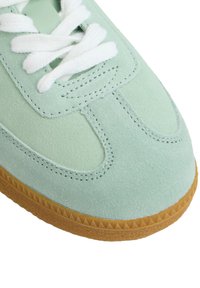 Zapatilla de ante verde claro con una superficie texturizada, cordones blancos y una suela de goma con un patrón de zigzag.