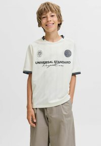 Niño sonriente con cabello rubio ondulado, vistiendo una camisa blanca de manga corta con texto negro y pantalones cortos beige, de pie frente a un fondo sencillo.