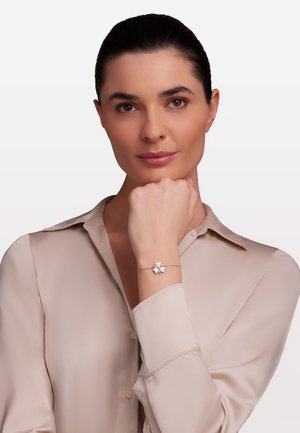 Goldarmband mit einem charmierenden weißen Blumenanhänger, vor einem hautfarbenen Hintergrund, getragen an einem Handgelenk eines Models. Glatte Bluse aus angenehm neutralem Stoff.