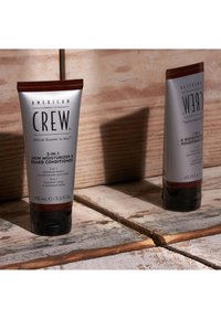 En tub av American Crew 2-i-1 hudfuktighetskräm och skäggbalsam. Silverfärgad kropp med brun kork, märkt med produktinformation i svart text.