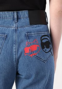 Blå denimjeans med svart lærbelte, to baklommer med røde og svarte tegneseriestil grafikker av en vaskebjørn og et ansikt med solbriller.
