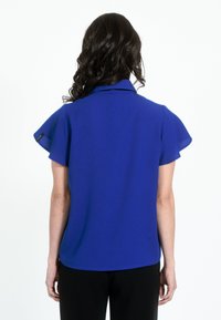 Kongeligt blå bluse med korte, svajende ærmer og kraveudskæring. Glat stof med en buet kant og diskrete branddetaljer.