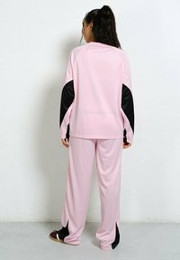 Kobieta stoi przy ścianie, ubrana w luźny różowy dres z czarnymi detalami na rękawach i nogawkach spodni, zwrócona plecami do aparatu.