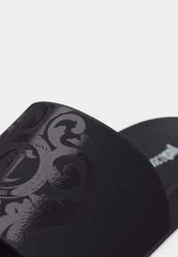 Μαύρη σαγιονάρα με γυαλιστερό, διακοσμητικό σχέδιο, με στυλιζαρισμένο γράμμα και υφή στην σόλα, σε απλό φόντο.