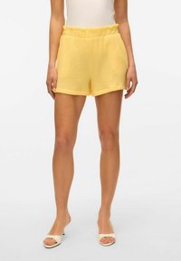 Żółte szorty wykonane z teksturowanego, lekkiego materiału, z marszczoną talią i luźnym krojem, w połączeniu z kremowymi klapkami na obcasie.
