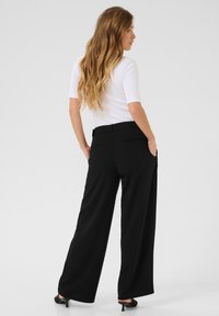 Pantalons noirs à jambe large en tissu lisse avec deux poches arrière. Associés à un haut blanc ajusté, mettant en valeur une silhouette propre et bien taillée.