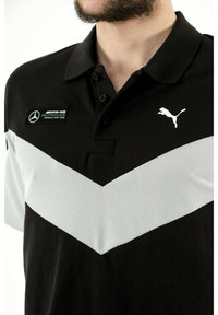 Zwarte poloshirt met een wit chevron ontwerp, wit Puma-logo en geborduurde badge van het Mercedes-AMG Petronas Formula One Team.
