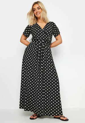 Kvinde med blondt hår iført en sort maxikjole med hvide prikker og korte ærmer, stående med hænderne bag ryggen og smilende.