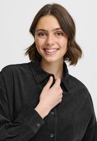 Camisa de pana negra con cuello, cierre de botones en el frente y mangas largas. Presenta una tela texturizada y un corte holgado.