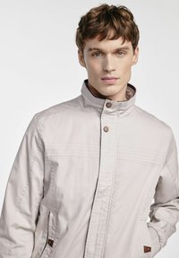 Jasno beżowa kurtka z kołnierzem stojącym, podwójnym zapięciem na guziki i dwoma bocznymi kieszeniami. Posiada fakturowany materiał i poziome przeszycia.