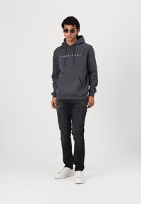 Sweat-shirt gris avec capuche et logo, jean noir et baskets blanches. Le modèle se tient avec les mains dans les poches, portant des lunettes de soleil rondes.